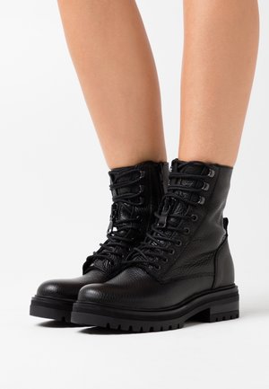 Schwarze gefütterte Leder-Kampfstiefel mit dicken Profilsohlen, Schnürsenkeln und seitlichen Reißverschlüssen, getragen an nackten Beinen vor weißem Hintergrund.