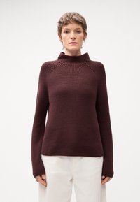 Maglione in borgogna lavorato a maglia con colletto alto a costine, maniche lunghe e tessuto testurizzato. Abbinato a pantaloni chiari.