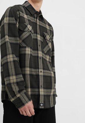 Mand iført sort og grå ternet skjorte med knapper og to brystlommer, stående mod en ensartet lys baggrund.