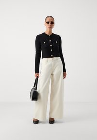 Black cropped sweater with gold buttons, paired with wide-leg ivory trousers. Model holds a black handbag and wears sleek sunglasses.