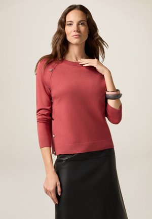 Roter Strickpullover mit Dreiviertelärmeln, ausgestattet mit Knopfdetails an der Schulter und einem gerippten Saum. Kombiniert mit einem schwarzen Lederminirock.