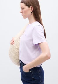 Ανοιχτό μωβ βαμβακερό μπλουζάκι με κοντά μανίκια και κεντημένο λογότυπο, σε συνδυασμό με σκούρο τζιν παντελόνι και ένα αφράτο, κρεμ τσαντάκι.