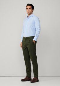 Cămașă albastru deschis, cu butoni și pantaloni verzi măslinii. Încălțămintea este bocanci din piele maro închis. Material neted, design fitted și stil clasic.
