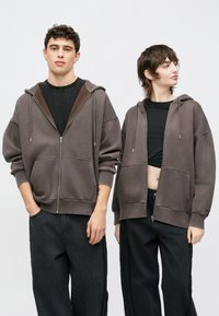 Δύο άτομα φορούν φαρδιές φούτερ με φερμουάρ σε απαλό γκρι χρώμα. Το ύφασμα είναι μαλακό με εμφανείς ραφές και μεγάλες μπροστινές τσέπες.