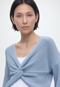 Sieviete ar īsiem tumšiem matiem, valkājot sudraboti sudraba auskarus un gaiši zilu adītu džemperi, kas pārklāts pār baltu t-kreklu, skatās uz priekšu.