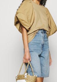 Hnědý top s nafouklými rukávy vyrobený z bavlny, ve spojení s modrými džíny s vysokým pasem. Modelka drží kulatou slaměnou tašku s bílými akcenty.