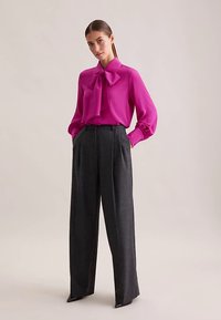 Camicia rosa acceso con un grande colletto a fiocco, abbinata a pantaloni color carbone a vita alta e taglio sartoriale, con pieghe marcate. Texture lisce.