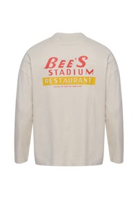 Bež majica z dolgimi rokavi z rdečim napisom "Bee's Stadium" in rumenim pasom "Restaurant" z manjšim sloganom na hrbtu.