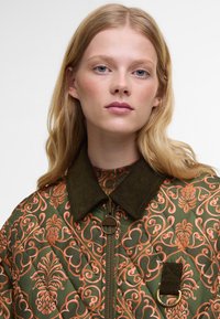 Casaco acolchoado verde com um intrincado padrão floral em laranja, apresentando um colarinho de sarja, fecho de zip e ferragens decorativas.