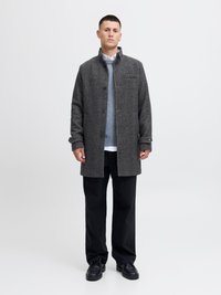 Cappotto grigio in lana con chiusura a bottoni, due tasche laterali e una vestibilità rilassata, indossato sopra un maglione azzurro chiaro e una camicia bianca con colletto. Pantaloni neri.