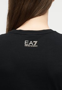 Άτομο με μακριά καστανά μαλλιά που φοράει μαύρο πουκάμισο με λευκό λογότυπο "EA7 Emporio Armani" τυπωμένο στο πάνω μέρος της πλάτης.