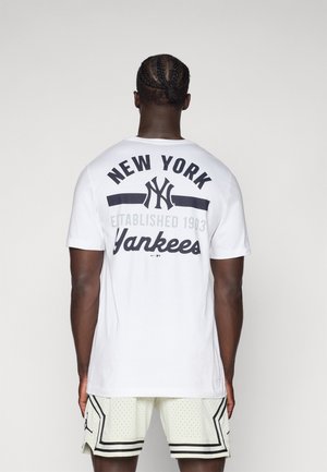 Mann mit geflochtenen Haaren, trägt ein weißes New York Yankees T-Shirt und weiße Basketballshorts mit schwarzen Akzenten, steht mit dem Rücken zum Betrachter.