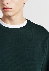Maglione verde scuro con texture, caratterizzato da un motivo a coste, scollatura rotonda con colletto bianco a contrasto, in primo piano sulla parte superiore del corpo.