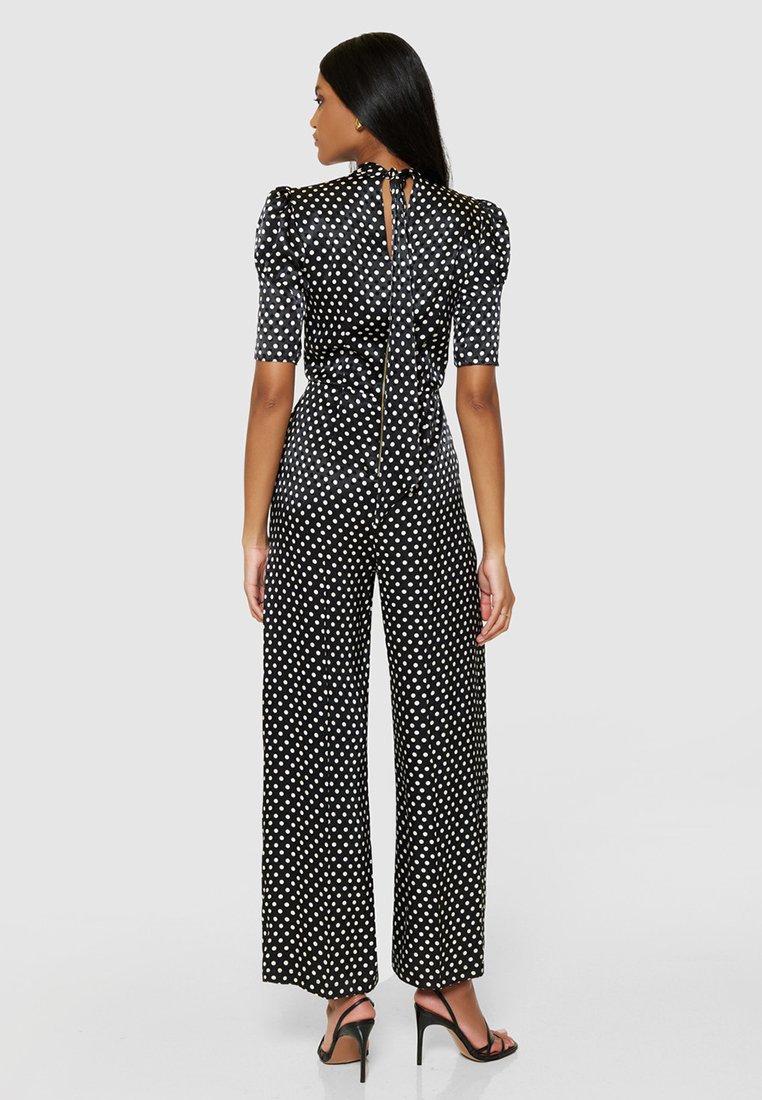 Czarny kombinezon w białe kropki z krótkimi bufkami, wysoką szyją, odkrytymi plecami z wiązaniem oraz szerokimi nogawkami. Gładka tekstura.