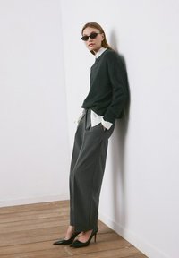 Mujer con suéter oscuro, camisa blanca, pantalones gris de pierna ancha, tacones negros y gafas de sol, apoyada contra una pared blanca sobre un suelo de madera.