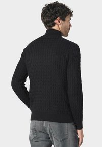 Czarny sweter dzianinowy z teksturowanym wzorem oraz ściągaczami przy kołnierzu, mankietach i dolnym brzegu. Noszony z szarymi dżinsami, prezentującymi dopasowaną sylwetkę.