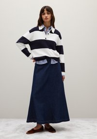 Gestreifter navy- und weißer Strickpullover mit Kragen, über einem blau- und weiß gestreiften Hemd getragen. Langer blauer Jeansrock und braune Slipper.