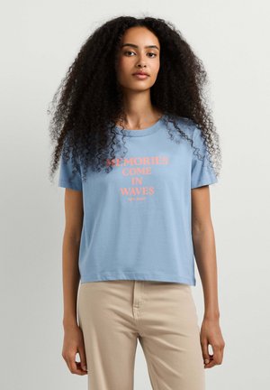 Femme aux longs cheveux bouclés portant un t-shirt bleu clair avec l'inscription « MEMORIES COME IN WAVES » et un pantalon beige sur un fond uni.