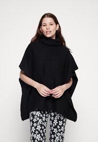 Svart strikket poncho med høy krage og løse ermer. Kombinert med bukser med svart-hvitt blomstermønster. Myk tekstur, oversized passform.