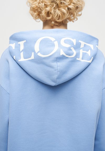 Άτομο με ξανθά σγουρά μαλλιά, ορατό από πίσω, φοράει γαλάζιο φούτερ με κουκούλα, στην οποία υπάρχει το λευκό κείμενο «CLOSE».