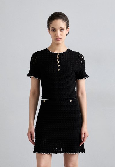 Schwarzes Strickkleid mit einem gewellten Ausschnitt, kurzen Ärmeln und Knopfakzenten. Mit weißem Saum und zwei Vorderentaschen.