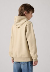 Felpa con cappuccio beige, dotata di un grande cappuccio, polsini e orlo a coste, realizzata in tessuto morbido. Indossata con jeans azzurri chiari. Visualizzata dalla parte posteriore.
