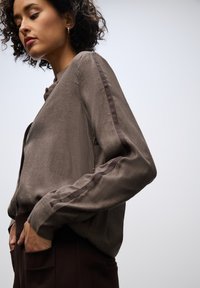 Blouse à manches longues marron en tissu texturé, avec fermeture à boutons sur le devant et rayures contrastantes marron foncé le long des manches.