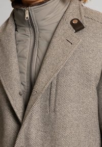 Grijze wolmix jas met een gestructureerde afwerking, met een hoge kraag, ritsluiting en bruine leren accentknopdetails.