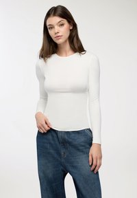 Giovane donna con capelli castani lisci, che indossa un top bianco a maniche lunghe attillato e jeans in denim blu larghi, su uno sfondo semplice.