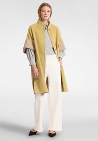 Capa abrigo mostaza con puños beige, con un alto cuello y dos botones. Combinado con una camisa a rayas y pantalones anchos blancos.
