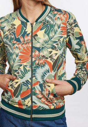 Femme portant une veste zippée avec un motif floral vert, orange et jaune, des poignets côtelés et un col rayé, mains dans les poches.