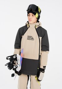 Giacca da neve beige e nera con cappuccio, caratterizzata da un logo, cerniere e dettagli gialli. Guanti neri e snowboard bianco con dettagli colorati.