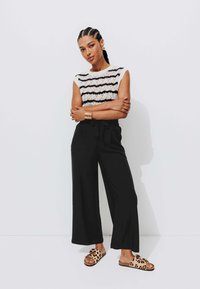 Pull en tricot avec motif à rayures noires, pantalon large noir, et sandales à semelle léopard marron. Manchette en laiton au poignet.