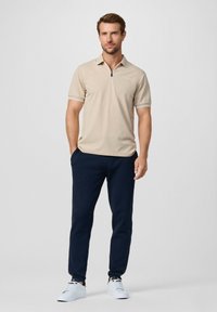 Polo beige a maniche corte con collo a zip, abbinata a pantaloni jogging blu navy e sneakers bianche. Design semplice con bordi a contrasto.
