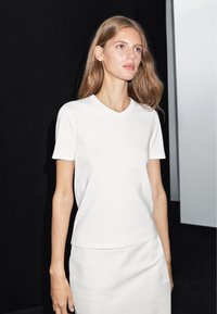 Biała, krótko rękawowa koszulka z dekoltem w serek. Wykonana z gładkiego, lekkiego materiału. Ma luźny krój i proste linie.