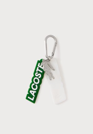 Srebrny karabińczyk z brelokiem trzyma zieloną zawieszkę Lacoste, srebrny urok krokodyla oraz białą teksturowaną zawieszkę.