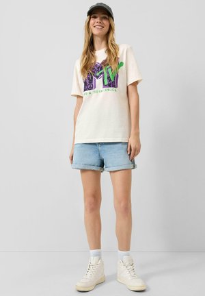 Junge Frau lächelt, trägt eine schwarze Kappe, weißes MTV Grafik-T-Shirt, hellblaue Jeansshorts, weiße Socken und weiße High-Top-Sneaker.