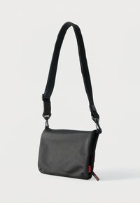 Μαύρη ορθογώνια τσάντα ώμου με ρυθμιζόμενο λουρί και κόκκινες ετικέτες λογότυπου στο πλάι, σε απλό λευκό φόντο.