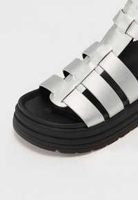 Sandalie din piele sintetică argintie cu patru curele orizontale, talpă groasă neagră și suprafață texturată. Dispune de un vârf rotunjit și un design minimal.