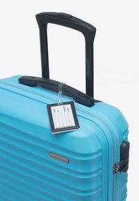 Tvrdý kufr v světle modré barvě s rýhovaným designem, černou zatahovací rukojetí, štítkem pro osobní údaje a kovovým logem značky.