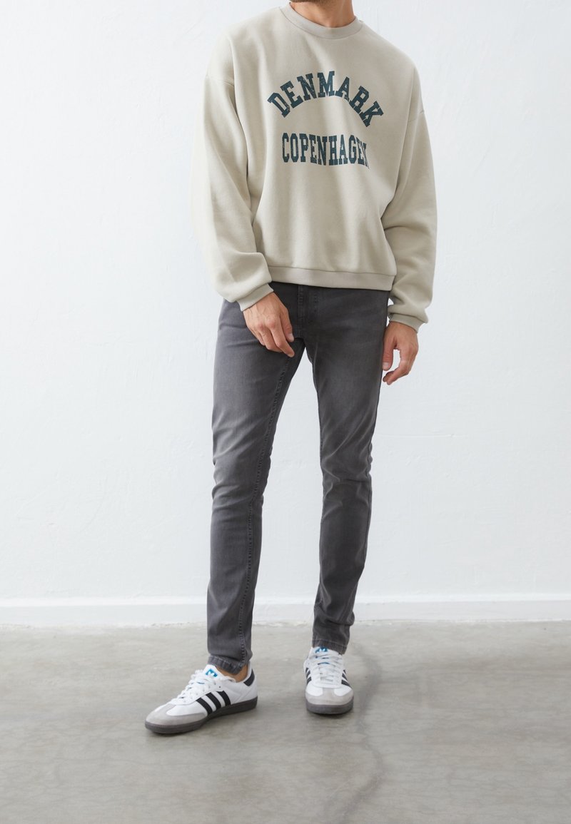 Sudadera de color gris claro con el texto "DINAMARCA COPENHAGUE", combinada con jeans gris oscuro y zapatillas blancas con rayas negras. Textura suave.