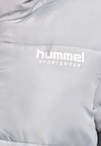 Lysgrå pufferjakke med en glat, skinnende tekstur. Har et synligt hvidt logo "hummel" og "SPORTSWEAR" på brystområdet.