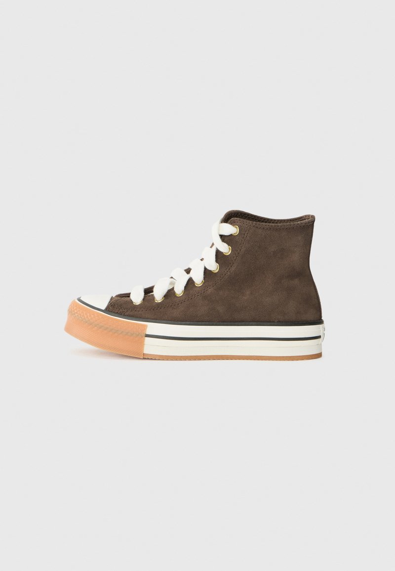 Zapatilla alta de ante marrón con cordones blancos, puntas de goma blanca y suela beige con rayas negras sobre un fondo claro.
