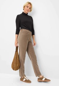 Schwarzer Rollkragenpullover, hellbraune Cordhosen, beige Sneaker mit weißen Akzenten und eine braune Umhängetasche mit Nieten. Glatte Texturen und figurbetontes Design.
