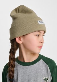 Olijfgroene gebreide beanie met een omslag, voorzien van een wit merkembleem. G gedragen met een grijs en groene raglan shirt, met gevlochten haar.