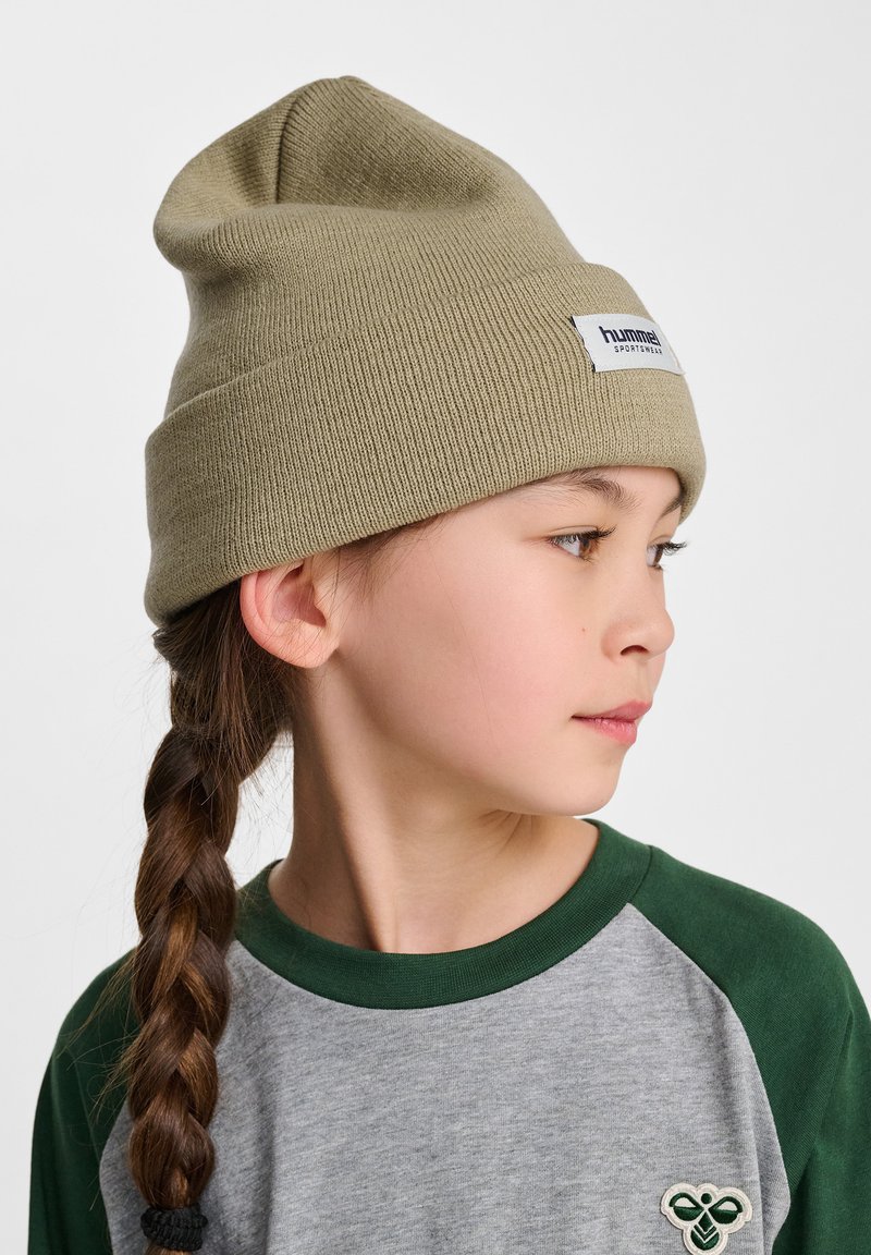 Olijfgroene gebreide beanie met een omslag, voorzien van een wit merkembleem. G gedragen met een grijs en groene raglan shirt, met gevlochten haar.