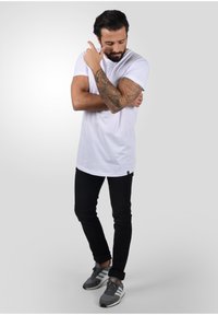 Camiseta blanca de algodón, jeans negros ajustados y zapatillas deportivas grises con rayas blancas. Tatuajes notables visibles en el brazo.