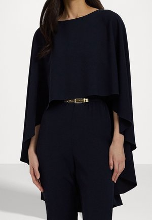 Kobieta ubrana w czarną bluzkę w stylu peleryny z szerokimi rękawami, połączoną z czarnymi spodniami z wysokim stanem i wąskim metalicznym paskiem.