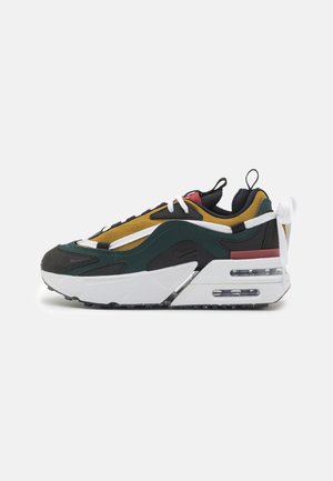 Zapatilla deportiva multicolor con suela blanca, superposiciones negras y verdes, parte superior de malla amarilla, detalles rojos y entresuela con amortiguación de aire visible.