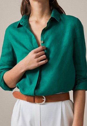 Kobieta ubrana w zieloną koszulę zapinaną na guziki z podwiniętymi rękawami, beżowe spodnie i brązowy pasek, zapinająca guzik koszuli prawą ręką.